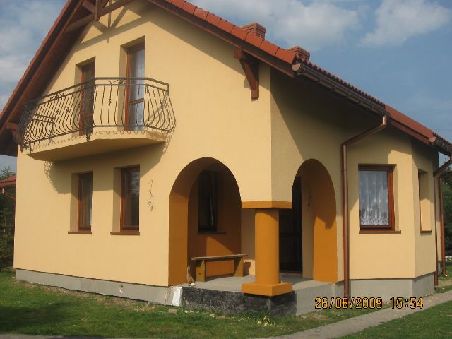 Żółty dom jednorodzinny z balkonem z kutą balustradą, łukowym wejściem i kolumnami w odcieniach pomarańczy, widok z zewnątrz.